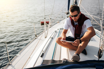 Serious and peaceful guy sits on board of yacht. He holds and looks at tablet. Young man is calm. He wears white sirt and dark sweater with shorts.