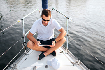 Young man sits with his legs crossed and hold tablet in hands. He is calm and concentrated. Screen is black. Guy sits on yacht bow. He wears sunglasses.