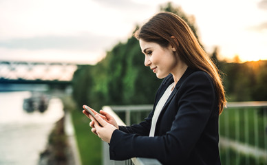 A young businesswoman standing on the river bank, using smartphone.