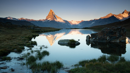 Sunrise at unspoilt Matterhorn with warm soft light on ridge and snow fields, clear blue sky, reflected on water Stellisee with stones and grass