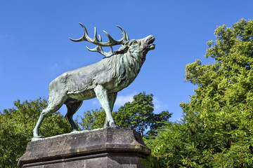 Big bronze statue of an antlered belling stag in a green park with trees and blue sky in the...