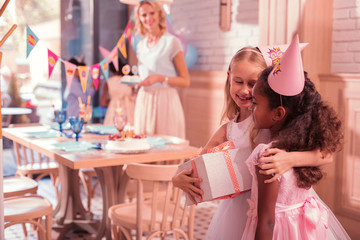Girl friendship. Pretty little girl holding a big present and smiling while kindly hugging her best friend