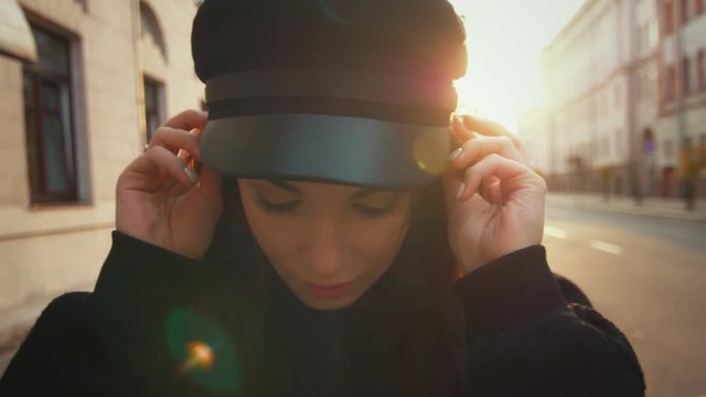 Pretty girl in urban landscape. Beautiful fashion model in black cap looking at camera. Woman walking in slow motion at sunset in street. Close up portrait of teenage girl