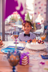 Relaxed boy. Positive relaxed boy wearing party hat and smiling while sitting at the table with big cake in front of him