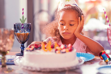 So boring. Gloomy dark haired girl leaning her head on her hand and thoughtfully looking at the cake while feeling tired