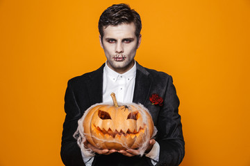 Photo of scary dead man on halloween wearing classical suit and creepy makeup holding carved pumpkin, isolated over yellow background