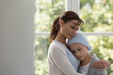 Sad mother hugging sick daughter with cancer wearing blue headscarf