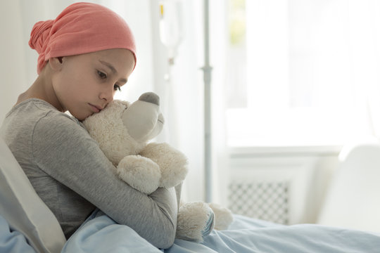 Weak girl with cancer wearing pink headscarf and hugging teddy bear