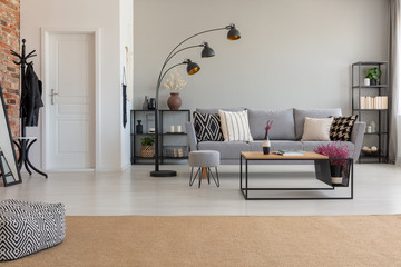 Pouf on carpet in spacious flat interior with lamp next to grey sofa and wooden table. Real photo