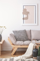 Poster above grey wooden settee with pillows in white flat interior with blanket and plant. Real photo