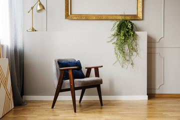 Gold lamp and plant above wooden armchair in grey flat interior with frame on the wall. Real photo