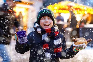 Little cute kid boy eating German sausage and drinking hot children punch on Christmas market - obrazy, fototapety, plakaty