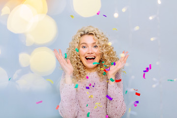Happy blond curly hipster girl enjoying and celebrating on a grey background. Blows up multicolored confetti. Photo taken through the garland light. Leisure, winter, holidays, people concept.