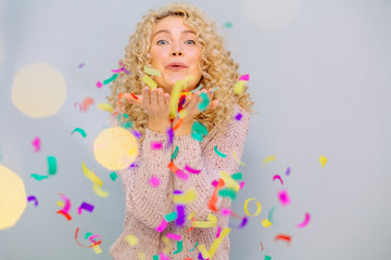 Obraz na płótnie Canvas Happy blond curly hipster girl celebrating on a grey background. Blows up multicolored confetti.