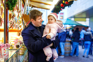 Obraz na płótnie Canvas Middle aged father holding baby daughter near sweet stand with gingerbread and nuts. Happy family on Christmas market in Germany. Cute girl eating cookie called Lebkuchen. Celebration xmas holiday.
