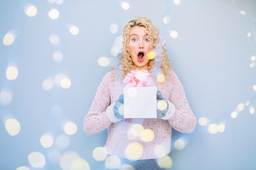 Schocked amazed curly blonde woman in knitted sweater and gloves with present box with pink bow indoor over gray wall background. Photo taken through the garland. Leisure, hygge, people concept.