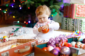 Adorable baby girl holding colorful vintage xmas toys and ball in cute hands. Little child in festive clothes decorating Christmas tree