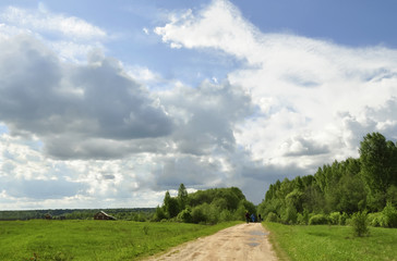 Obraz premium Summer landscape with meadow, wooden house far aside, blue sky, white clouds, three people going away on the rural dirt road with green grass by its both sides towards the woods afar in the horizon.