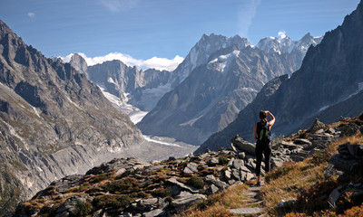 Naklejka na ściany i meble A lone female Hiker looks out over a glacier valley in the region of Chamonix in the alps of France. High alpine snow covered mountains surround the foreground rocks and alpine terrain.
