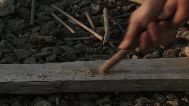 Young man making fire with wood stick on lake beach during sunset