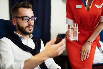 Portrait of young handsome man using smartphone with flight attendant bringing him drinks in background, copy space