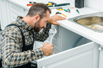 bearded repairman holding adjustable wrench and looking under broken sink in kitchen