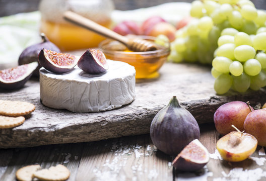 The set of natural products - soft cheese, figs, grape, plums, honey with stick on a wooden board.
