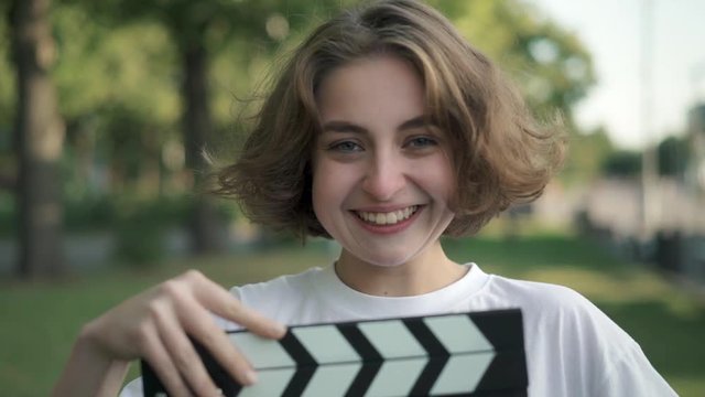 Serious young woman with short thick hair looking at camera. She is using clapperboard and starts laughing after that. Concept of filmmaking and acting. Slider slow motion portrait shot