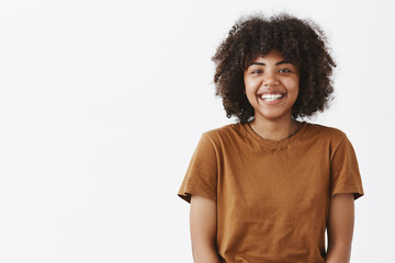 Waist-up shot of cute carefree friendly-looking African American teenage girl with afro hairstyle smiling broadly with shy and happy expression meeting new classmates over gray background - obrazy, fototapety, plakaty