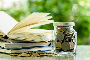 Money coins in the glass jar with stack of opened books against blurred natural green background...