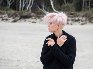 Outdoor lifestyle fashion portrait of amazing sexy young smoking woman with stylish, trendy haircut and pink hair, in a black cardigan, punk, hipster posing with cigarette