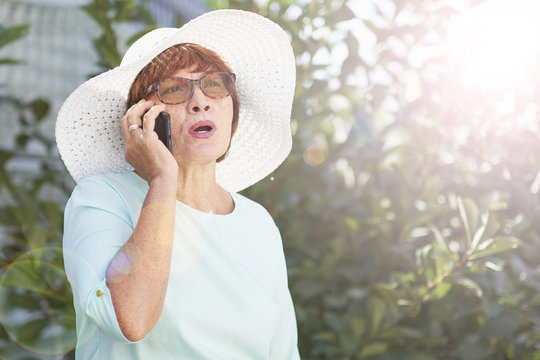 Aged Woman Talking On The Phone. Old Lady In Park. Sunny Day, Happy Mood. Upset Senior Woman With Phone