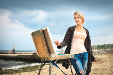 Beautiful young woman painting on the background of the sea and sky. The concept of creativity.