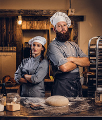 Chef with his assistant in cook uniform posing with crossed arms near table with ready dough in the...