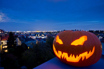 Halloween pumpkin face creates scary evening mood. Clear view to the cityscape of Vienna from the rooftops of the suburbs with city lights in the background.