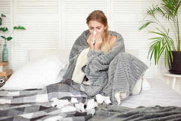 Miserable young woman sitting on the bed wrapped in warm blanket feeling sick with flu.