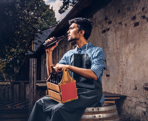 Brewmaster sitting on a wooden barrel and holds a glass of craft beer, relaxes after work.
