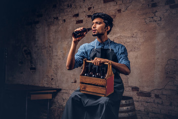 Brewmaster sitting on a wooden barrel and holds a glass of craft beer, relaxes after work.