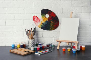 Naklejka premium Easel with various artist tools on table against brick wall. Space for text