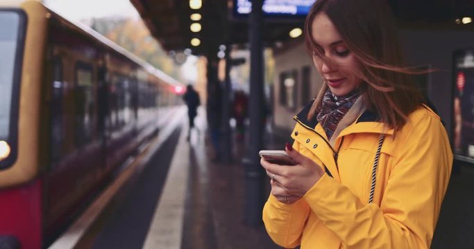 Young Woman Using Smartphone at the Train Station. SLOW MOTION 4K. Girl using cell phone, waiting for city train. Social network, planning, communicating.