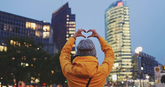 Young Woman Showing Heart Sign With Her Hands, Night Busy City Background. SLOW MOTION 4K. Unrecognizable Girl Loving big city life. Skyscrapers and traffic light at dusk.