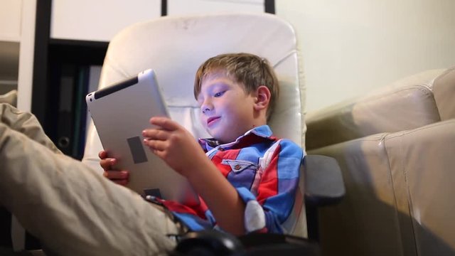 Happy young boy playing on his tablet sitting on the chair at home.