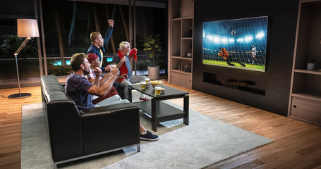 Group of fans are watching a soccer moment on the TV and celebrating a goal, sitting on the couch in the living room.