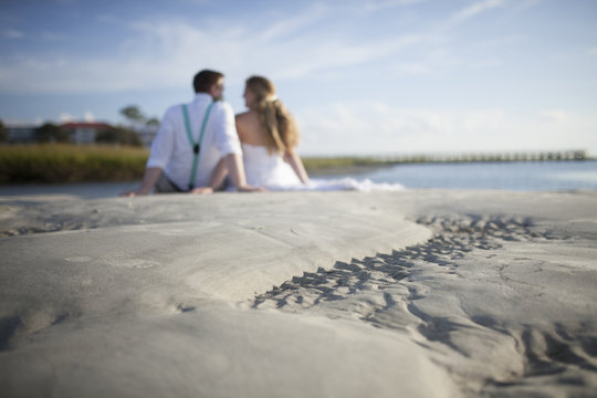 Newly married couple seated on beach, out of focus in background with focus on ripples in sand