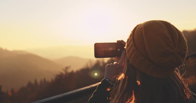 Young Woman Taking Pictures of Sunlit Fall Mountain Landscape form the Top. SLOW MOTION. Hiker Girl is Taking photos with smartphone of autumn hills landscape at sunset, Lens Flare.  