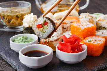 Two chopsticks holding Hosomaki roll with vegetables and different sushi rolls with seafood