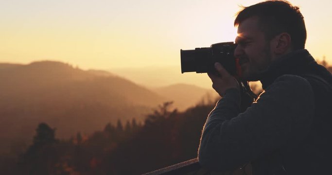 Young Man Taking Pictures of Sunlit Fall Mountain Landscape form the Top. SLOW MOTION. Travel Photographer is taking photos with digital camera of autumn hills landscape at sunset, Lens Flare.  