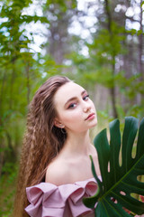 Naklejka premium a young teenager girl portrait with long curled hair dressed in pink in the forest with an exotic plant leaf