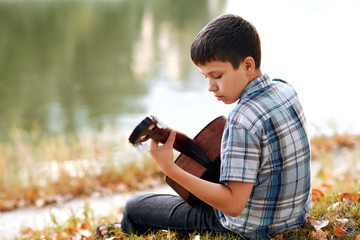 the boy plays an acoustic guitar, sits on the Bank of the river, autumn forest at sunset, beautiful nature and the reflection of trees in the water