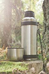 Fototapeta na wymiar Steel thermos with a cup of hot coffee standing on old stump in coniferous forest, camping abstract background